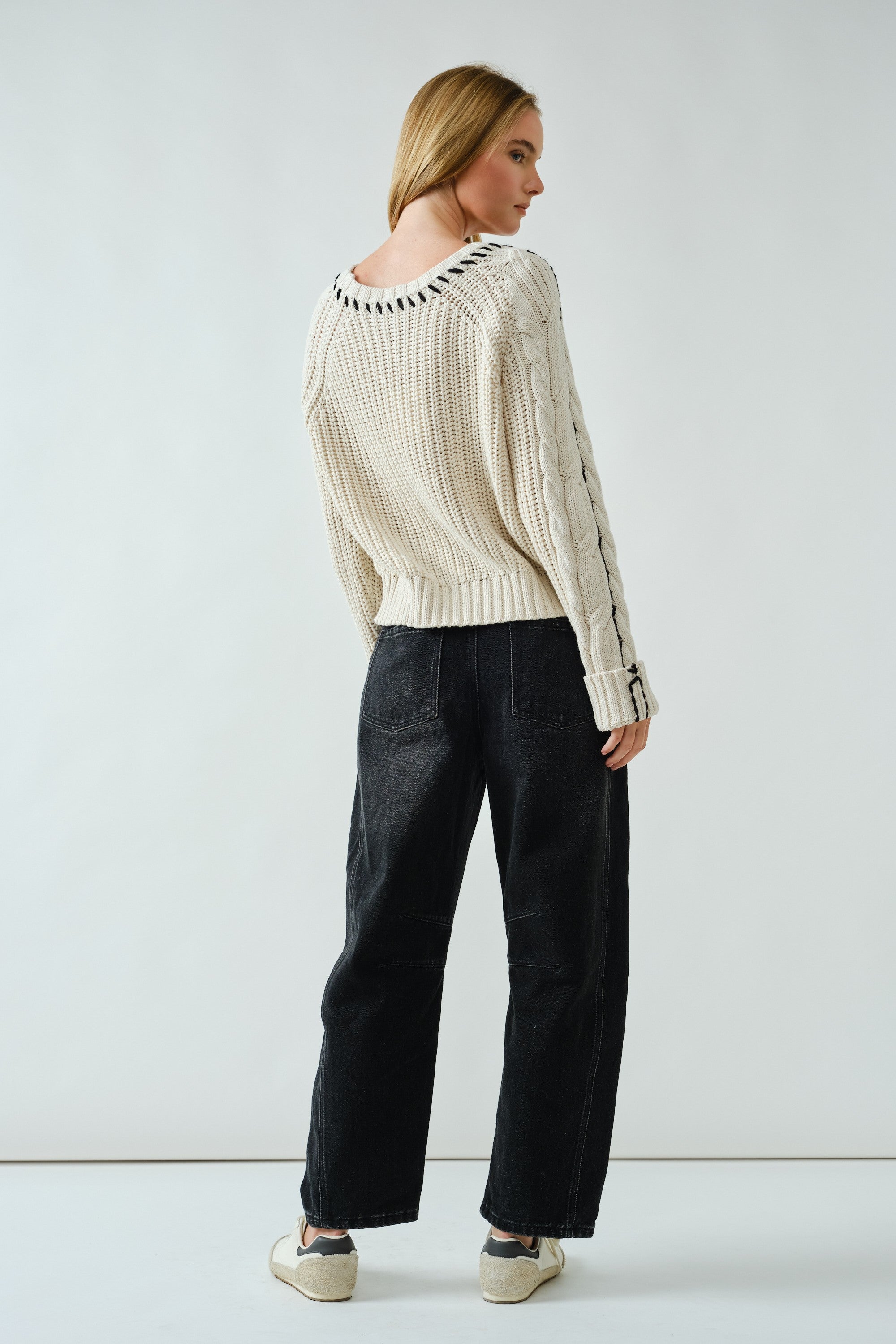 Woman wearing a cream sweater with black trim and dark pants on a white background