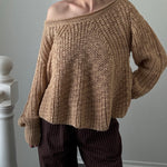 Woman wearing a brown knitted sweater and dark pants against a plain wall.