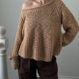 Woman wearing a brown knitted sweater and dark pants against a plain wall.