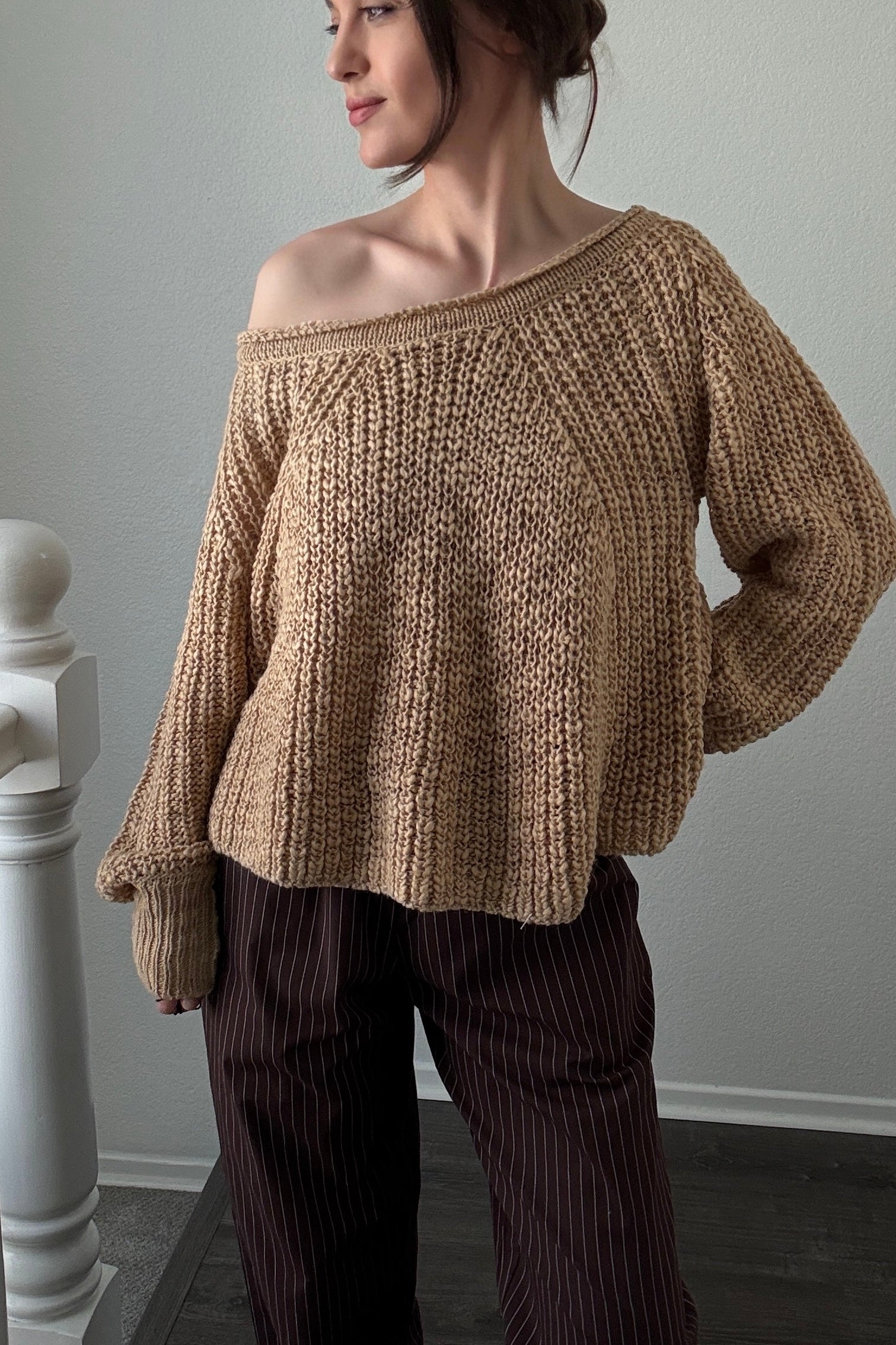 Woman wearing a brown knitted sweater and dark pants against a plain wall.