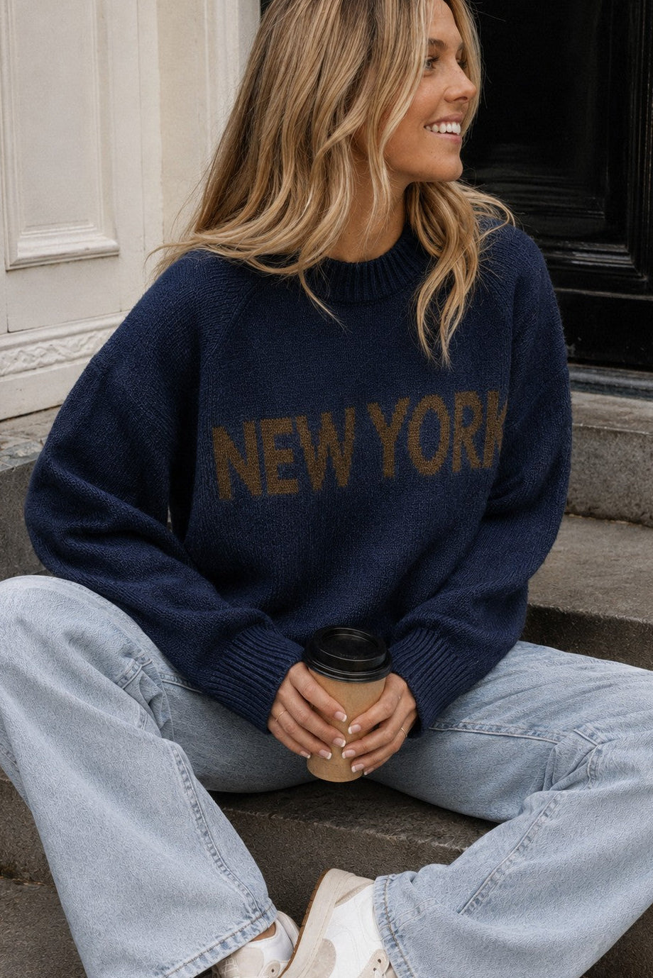Lifestyle image of woman wearing New York Graphic Sweater in Navy with bold NEW YORK lettering and light-wash jeans