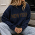 Lifestyle image of woman wearing New York Graphic Sweater in Navy with bold NEW YORK lettering and light-wash jeans