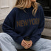 Lifestyle image of woman wearing New York Graphic Sweater in Navy with bold NEW YORK lettering and light-wash jeans