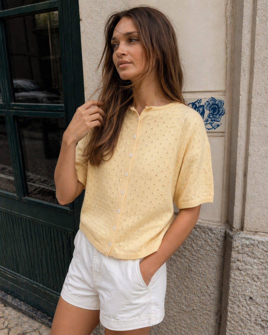 Woman wearing Piper Pointelle Cotton Button Down Cardigan - Light Yellow and white shorts standing against a textured wall.