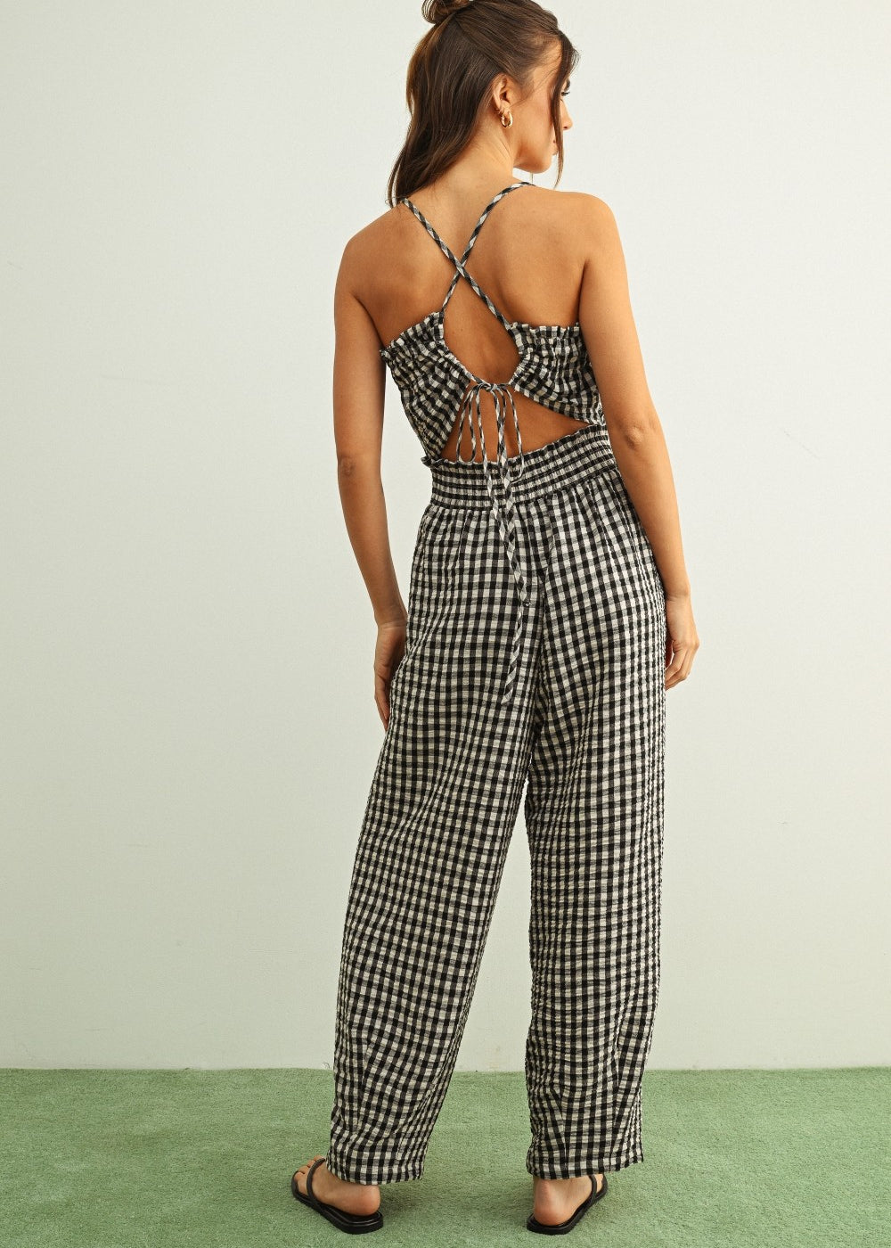 Woman wearing Black and white gingham smocked jumpsuit with barrel-leg fit and lightweight cotton fabric with cut-out back design on a light green background.