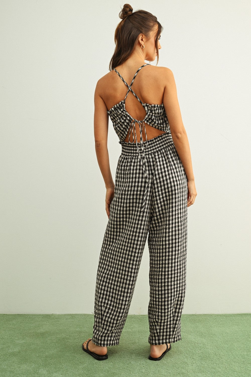 Woman wearing Black and white gingham smocked jumpsuit with barrel-leg fit and lightweight cotton fabric with cut-out back design on a light green background.