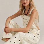 Woman wearing a beige sleeveless top and polka dot overalls sitting on a light gray background
