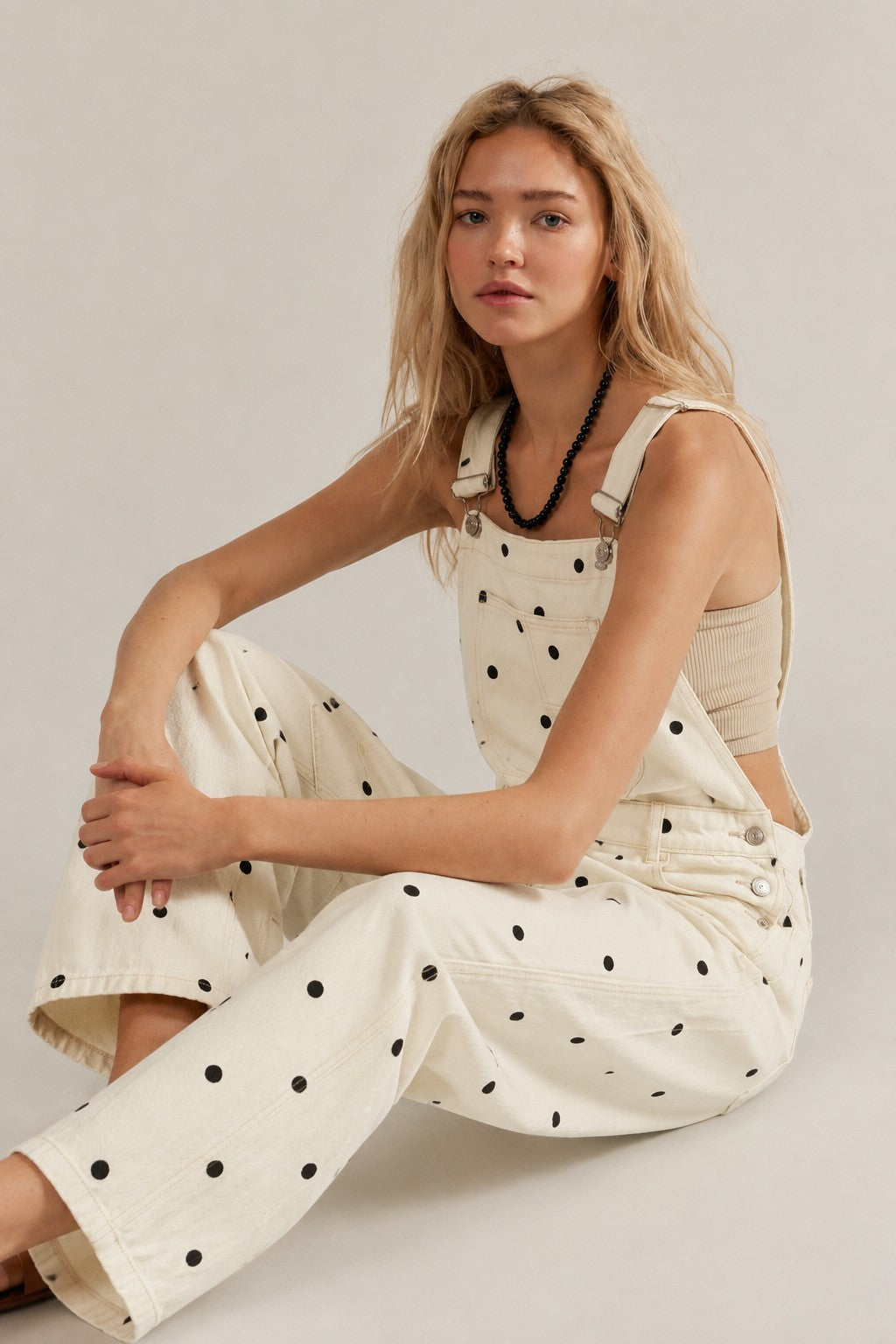 Woman wearing a beige sleeveless top and polka dot overalls sitting on a light gray background