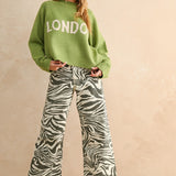 Woman wearing a green sweater with 'LONDON' text and zebra print pants on a beige background