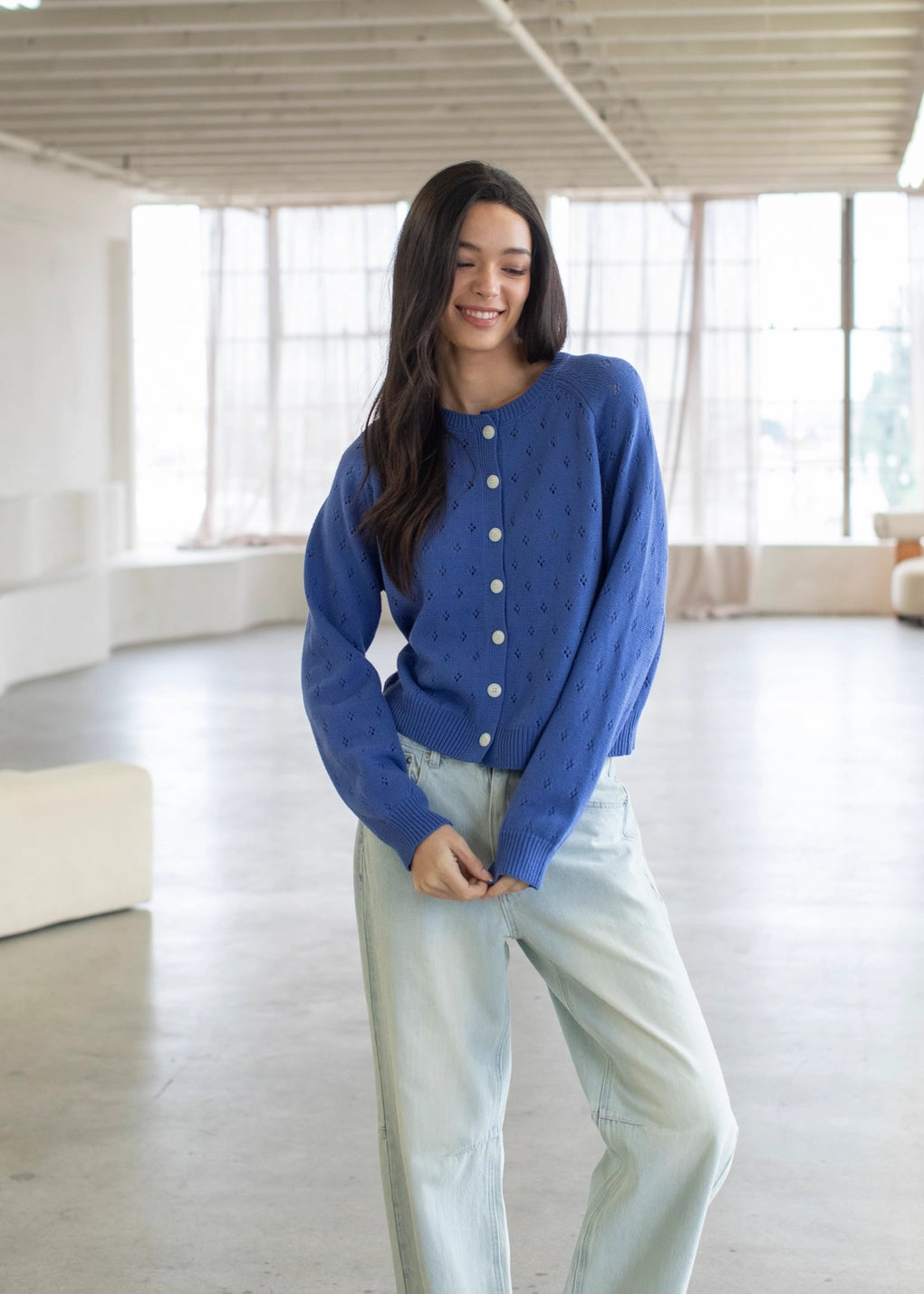 Woman wearing a blue cardigan and light jeans in a bright room with large windows.