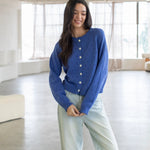 Woman wearing a blue cardigan and light jeans in a bright room with large windows.