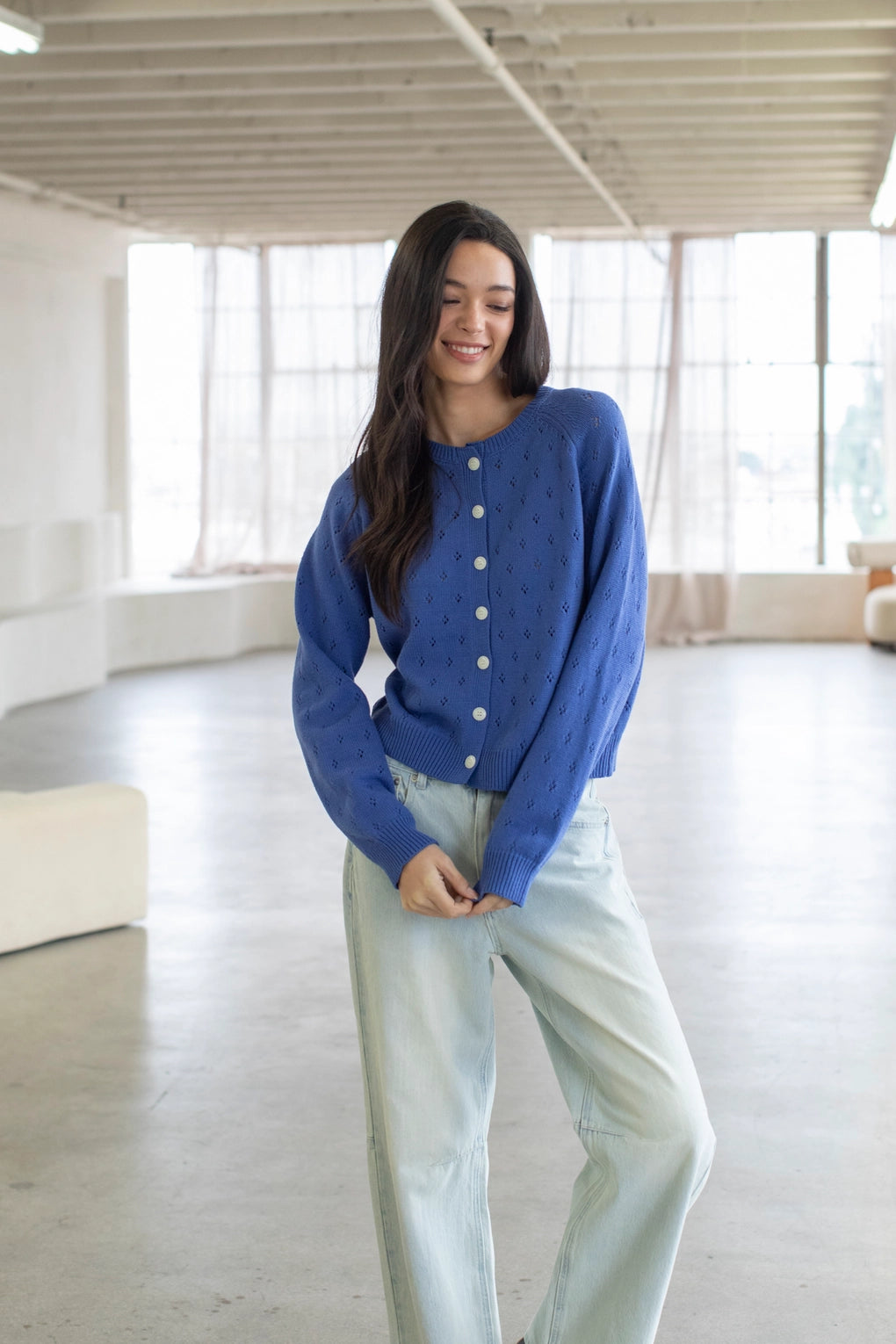 Woman wearing a blue cardigan and light jeans in a bright room with large windows.