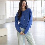 Woman wearing a blue cardigan and light jeans in a bright room with large windows.