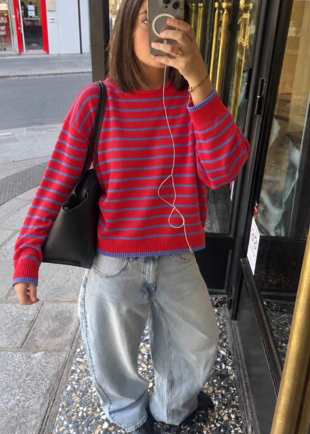 A woman takes a mirror selfie wearing a red and blue striped knit sweater, light-wash wide-leg jeans, and black shoes, carrying a black shoulder bag.