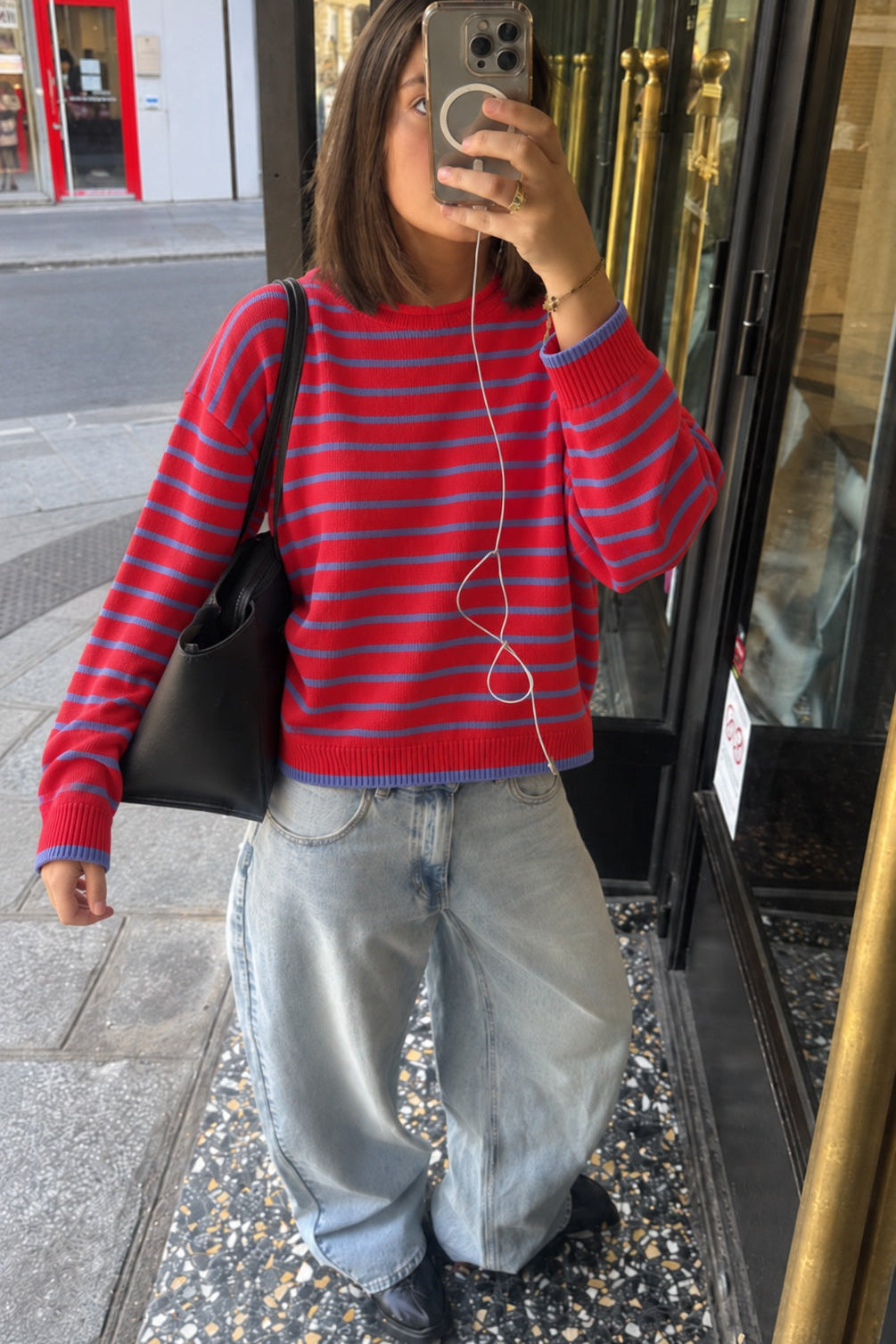 A woman takes a mirror selfie wearing a red and blue striped knit sweater, light-wash wide-leg jeans, and black shoes, carrying a black shoulder bag.