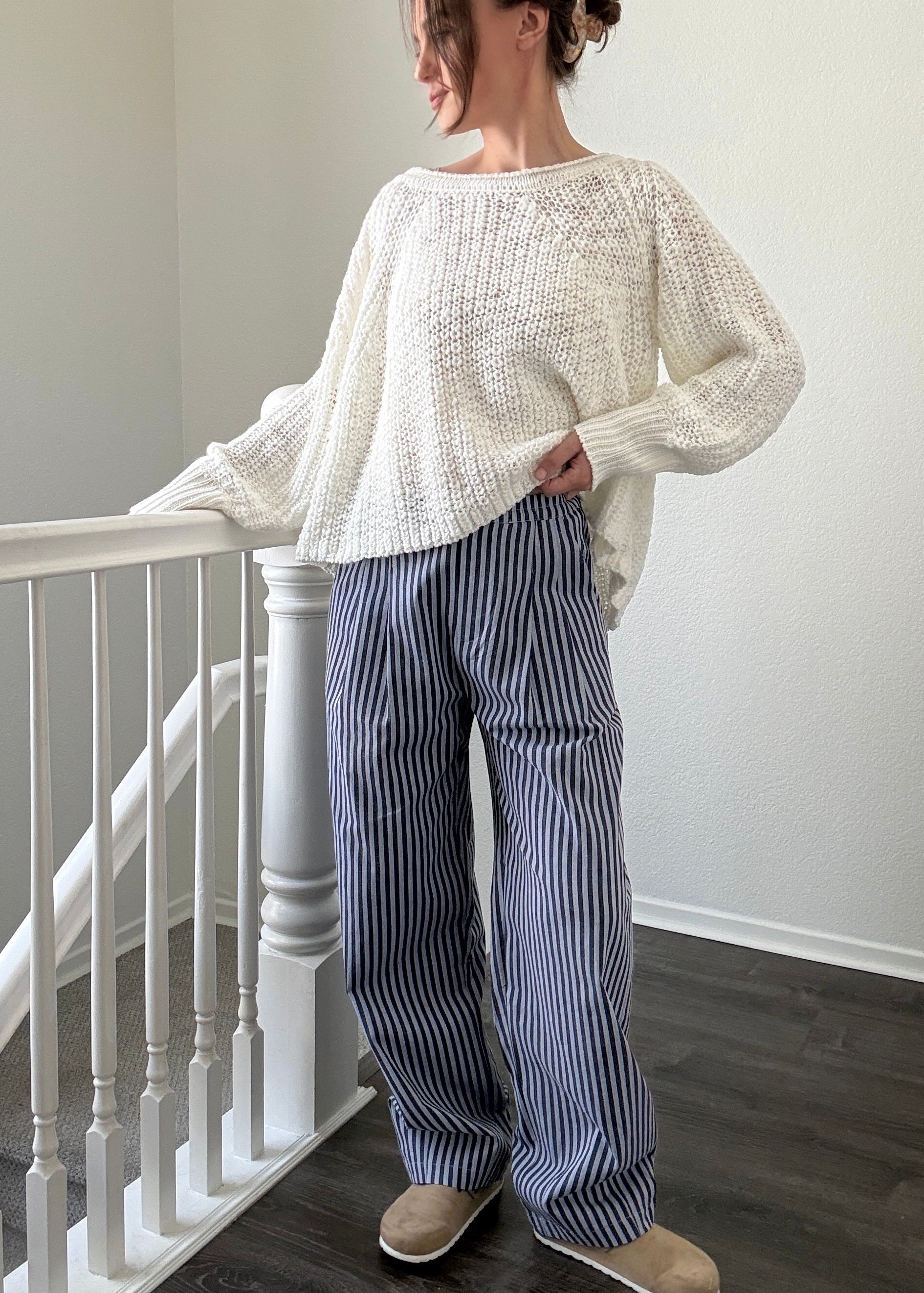 Person wearing a white sweater and blue striped pants standing in a room with a staircase.