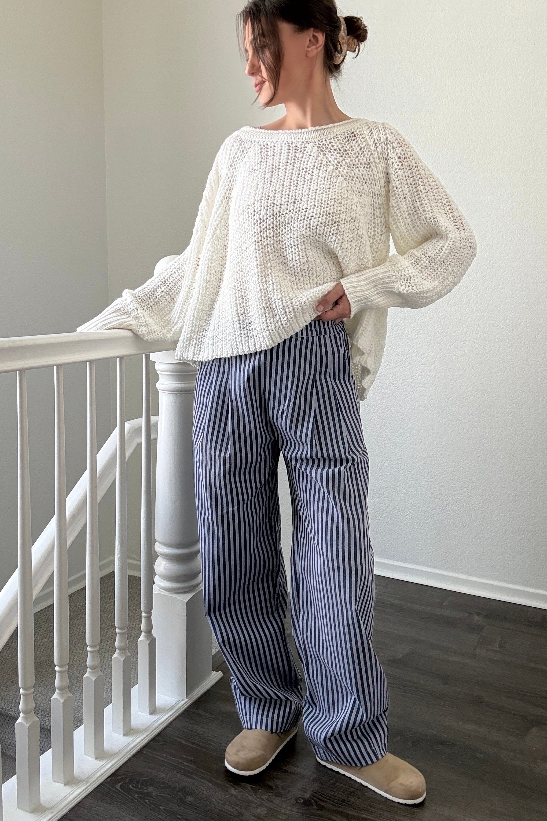 Person wearing a white sweater and blue striped pants standing in a room with a staircase.