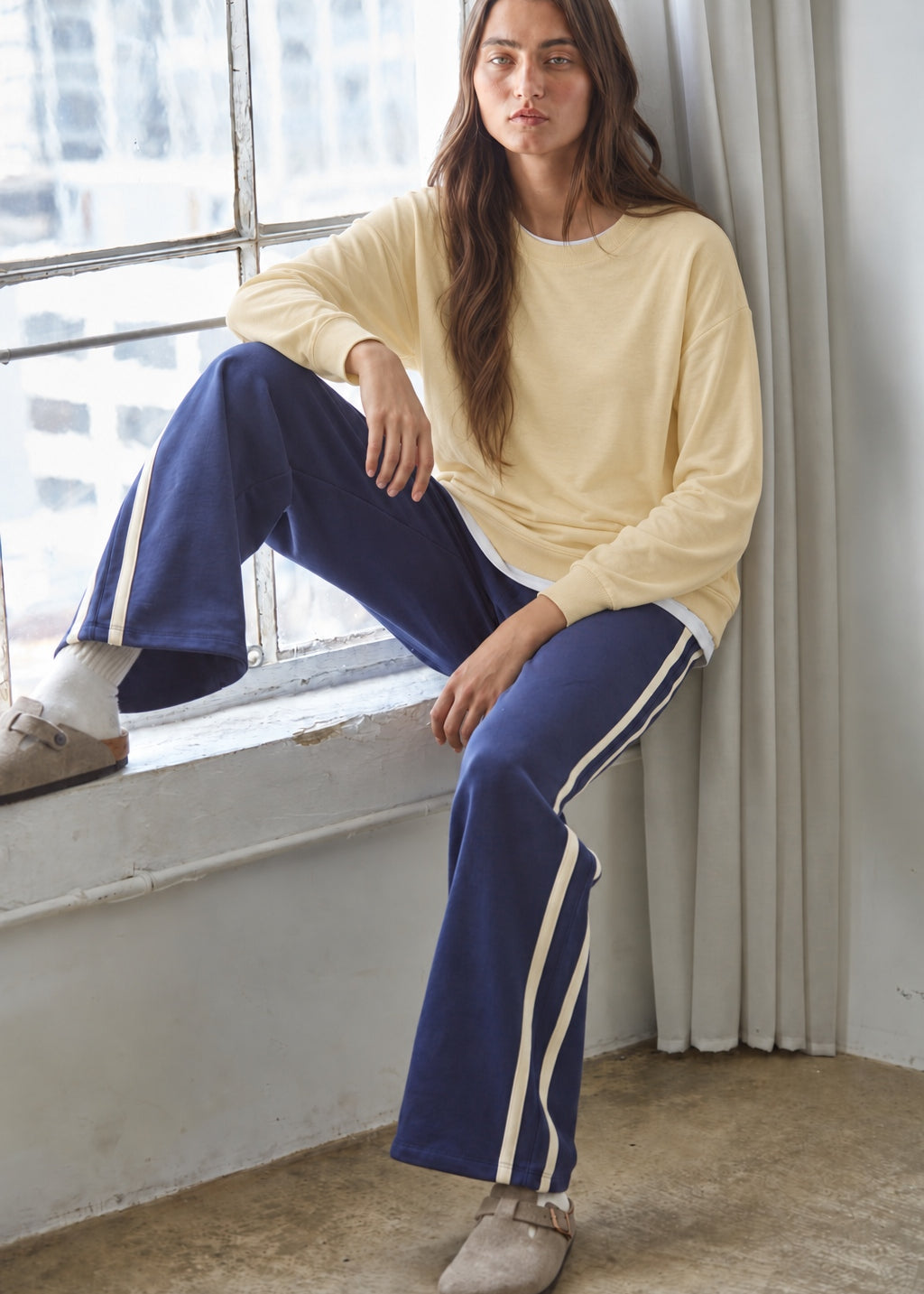 Woman seated beside a window wearing the Jetset butter yellow twofer sweatshirt with blue pants.