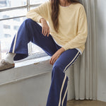 Woman seated beside a window wearing the Jetset butter yellow twofer sweatshirt with blue pants.