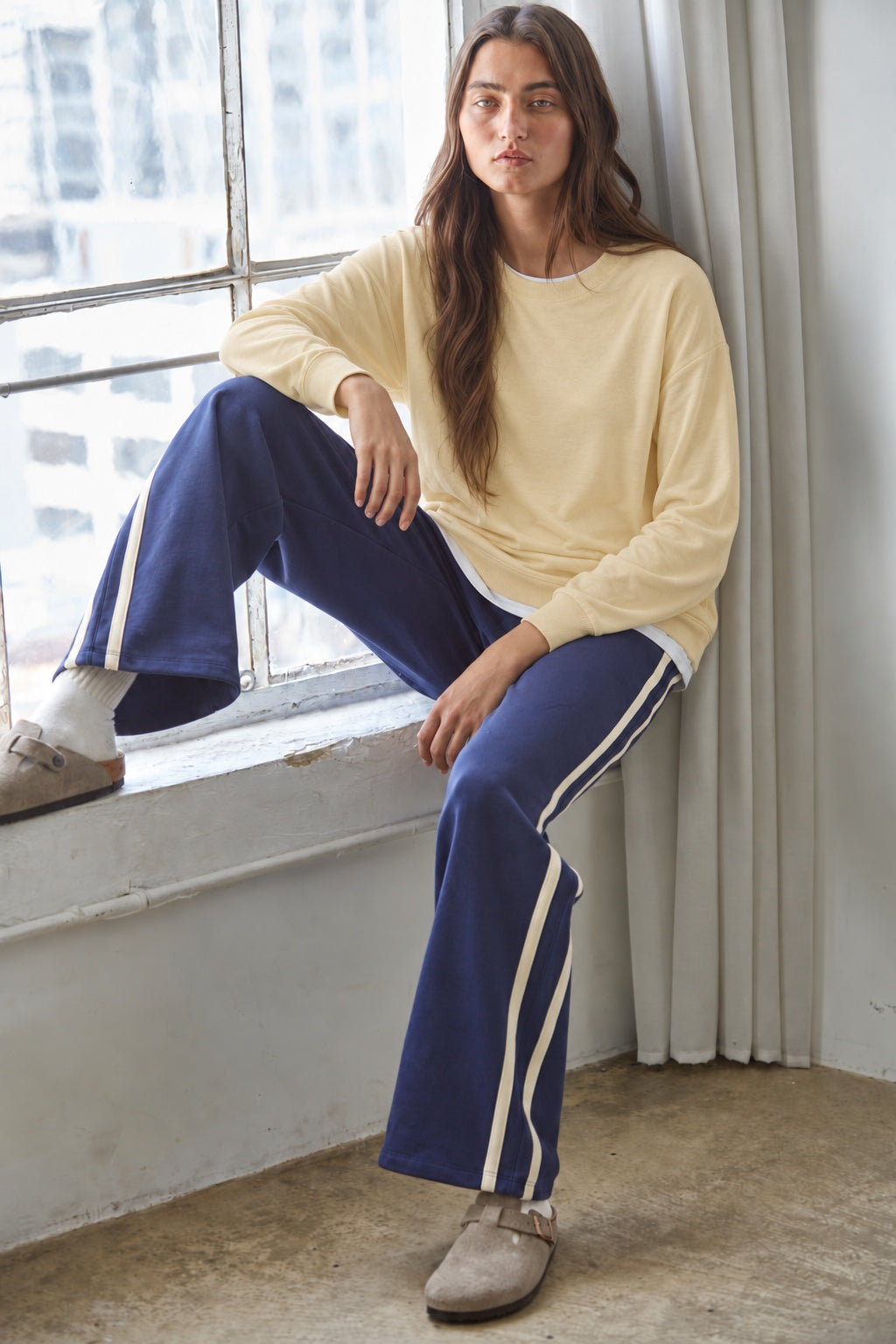 Woman seated beside a window wearing the Jetset butter yellow twofer sweatshirt with blue pants.