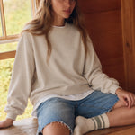 Woman seated beside a window wearing the Jetset cream twofer sweatshirt with denim shorts.