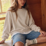 Woman seated beside a window wearing the Jetset cream twofer sweatshirt with denim shorts.