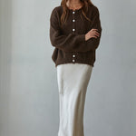 Woman wearing a brown cardigan and white skirt against a plain background