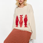 Woman sitting while wearing the Marina Fish Knit Sweater — soft sand knit with fun red fish design.
