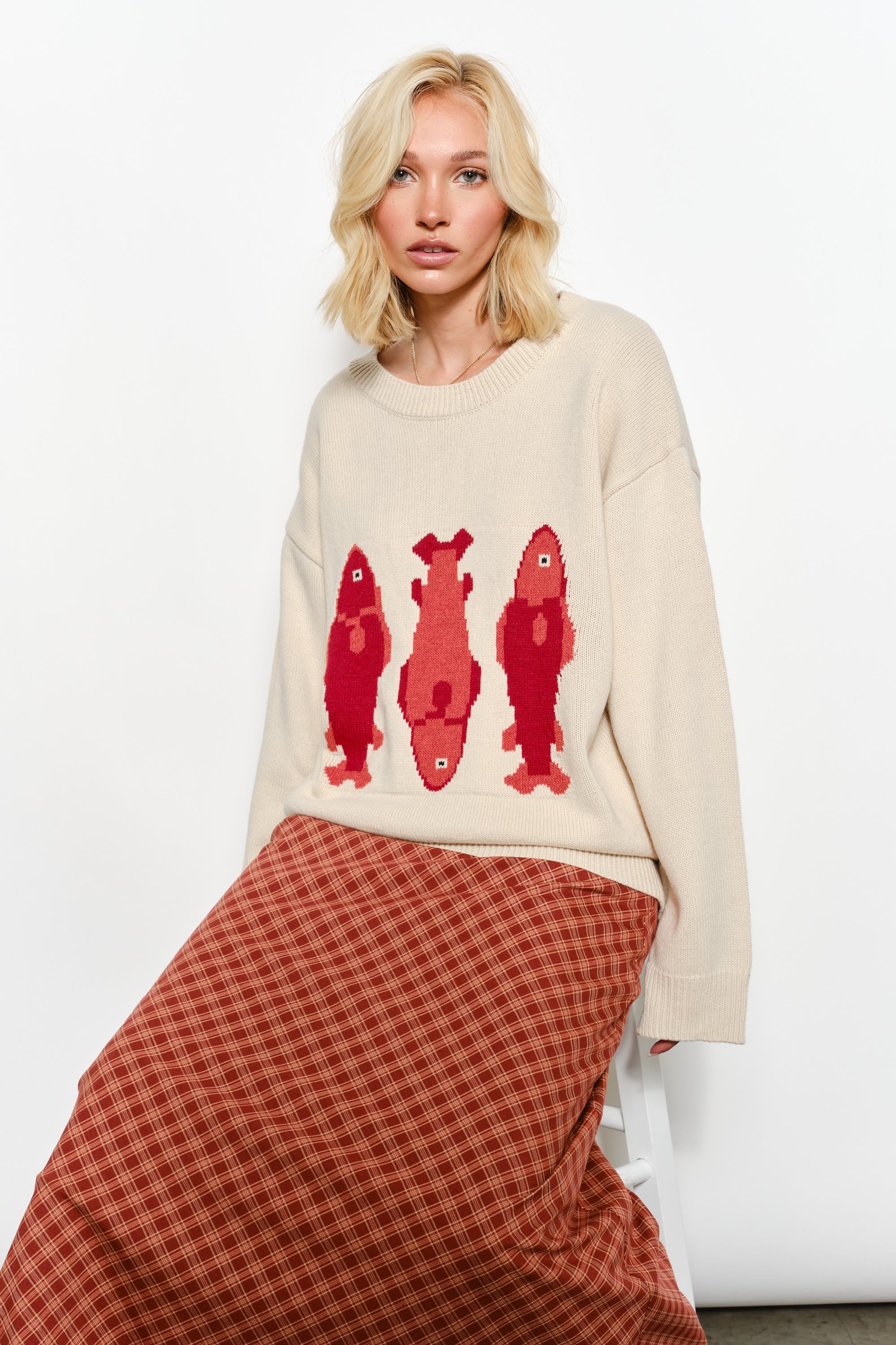 Woman sitting while wearing the Marina Fish Knit Sweater — soft sand knit with fun red fish design.
