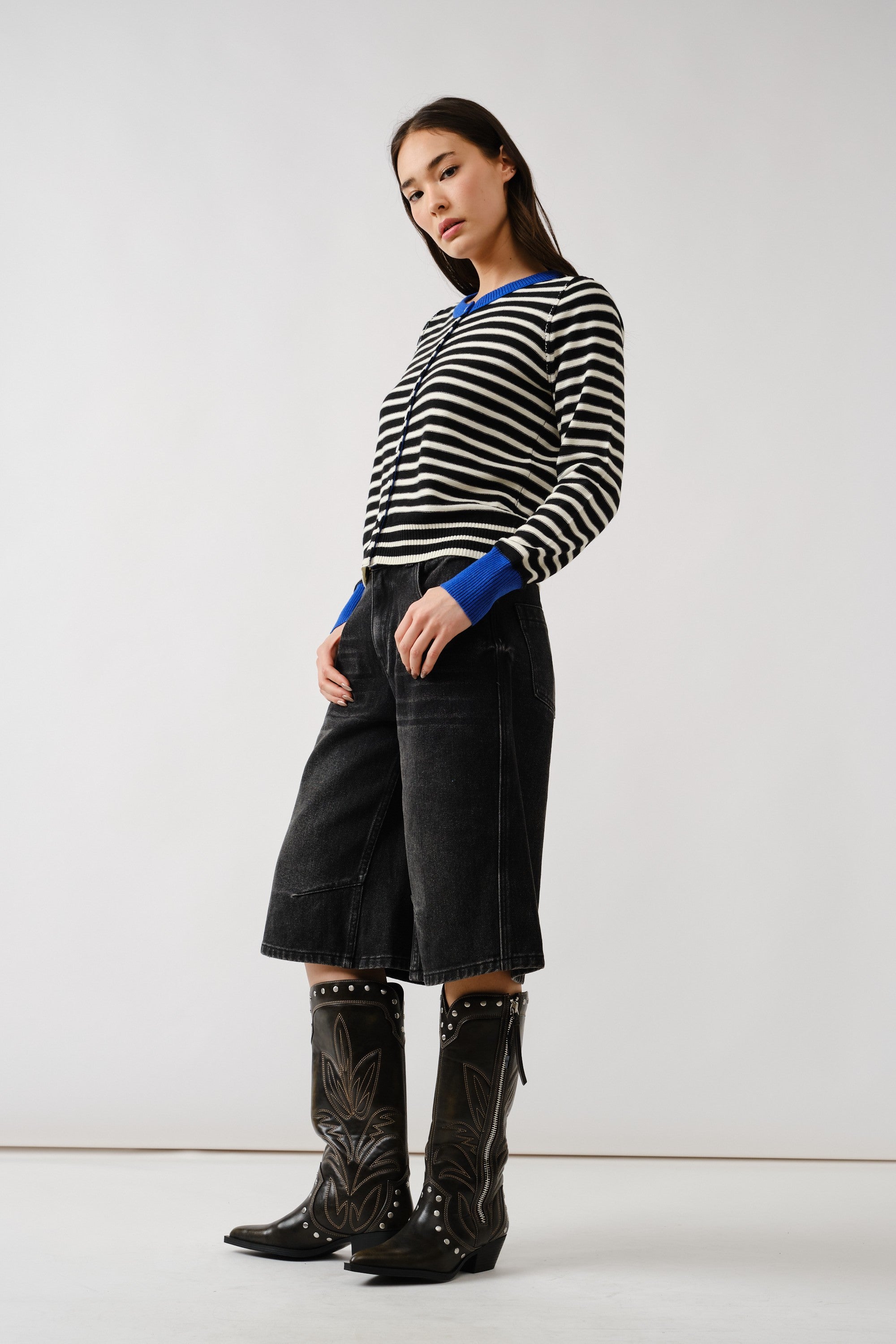 Woman wearing a black and white striped sweater with blue accents, black high-waisted shorts, and black cowboy boots on a white background.