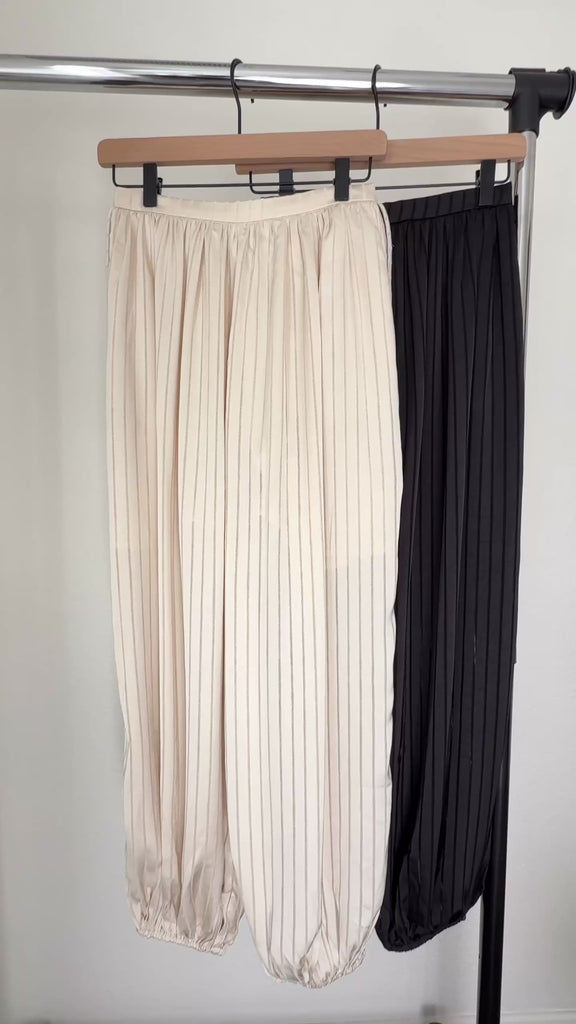 Close-up video showing the striped satin texture, elastic waistband, side drawstrings, pockets and balloon-leg shape of the beige Double Bubble pants.