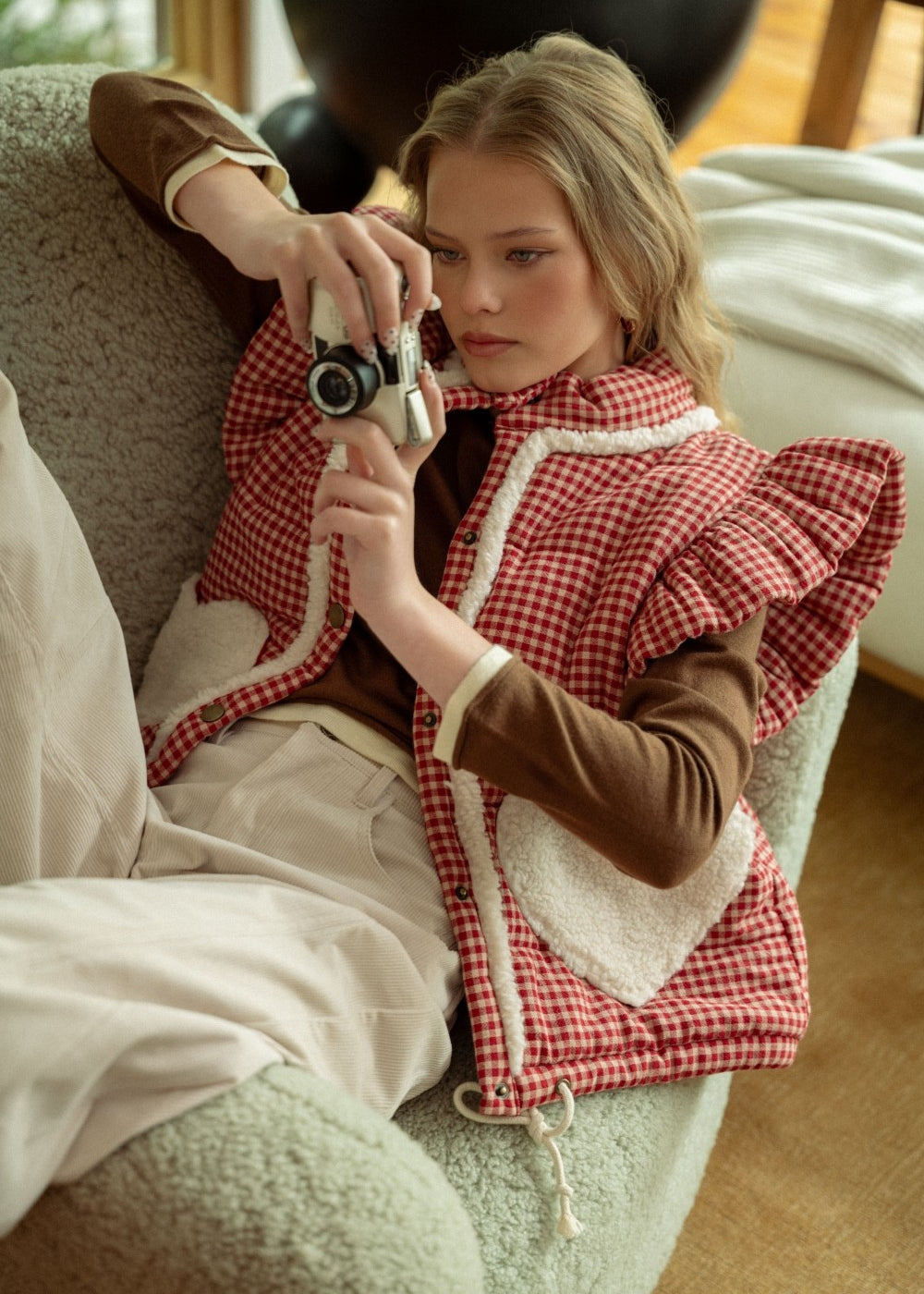 Lifestyle photo of model sitting on a couch wearing the red gingham sherpa puffer vest layered over a long sleeve top