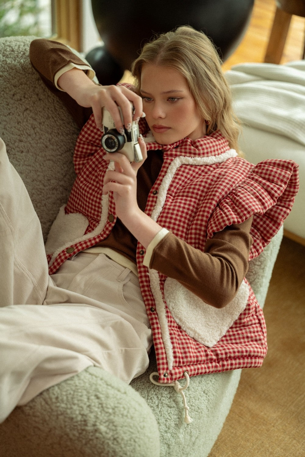 Lifestyle photo of model sitting on a couch wearing the red gingham sherpa puffer vest layered over a long sleeve top