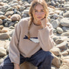 Lifestyle image of model wearing the Sardine Balloon Sleeve Sweater in oat, seated on coastal rocks