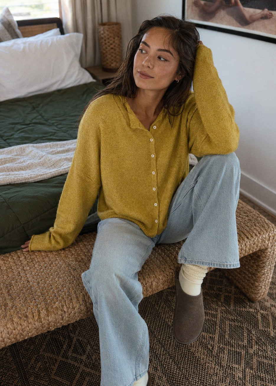 Woman wearing the Things Between Piper Cardigan in Dijon Yellow in a relaxed seated lifestyle setting