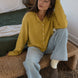 Woman wearing the Things Between Piper Cardigan in Dijon Yellow in a relaxed seated lifestyle setting