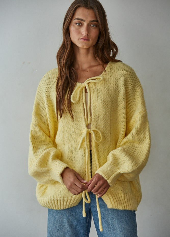Woman wearing a yellow knitted bow tie cardigan against a plain background
