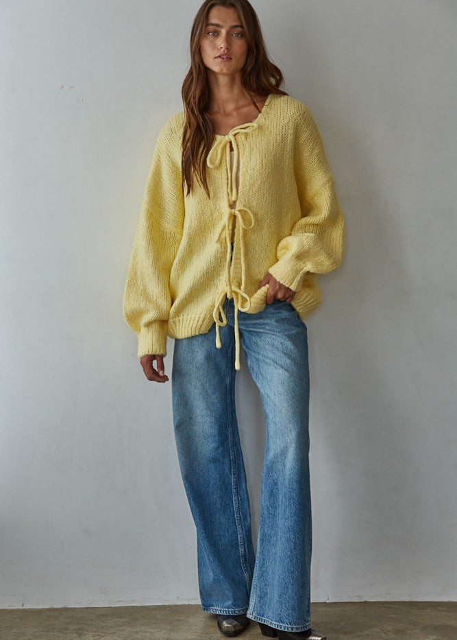 Woman wearing a yellow bow tie cardigan  and blue jeans against a plain background